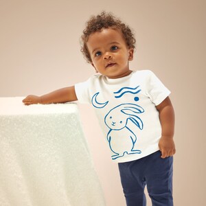 Może przedstawiać: Biała koszulka z niebieskim królikiem, księżycem i falistymi liniami. Koszulkę nosi małe dziecko z brązowymi włosami.