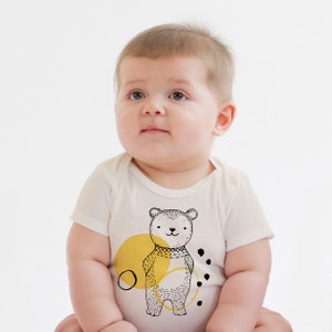 Könnte beinhalten: Ein weißer Baby-Strampler mit einer schwarz-weißen Illustration eines Bären, der einen gelben Schal trägt. Der Bär steht vor einem gelben Kreis mit schwarzen Punkten drum herum.