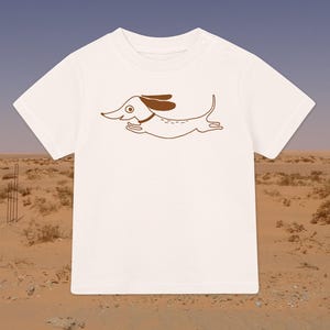 Może przedstawiać: Biała koszulka dla niemowlęcia z brązowym konturem biegnącego jamnika. Jamnik ma brązowe uszy i brązowy kołnierz.