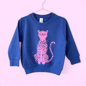 Può includere: Una felpa blu navy con un disegno di stampa leopardata rosa. La parte superiore a maniche lunghe è appesa a una gruccia di metallo su uno sfondo rosa chiaro. Il leopardo è seduto.