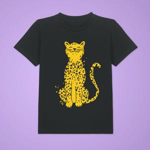 Może przedstawiać: Czarny t-shirt z żółtą ilustracją lamparta. Lampart jest przedstawiony w pozycji siedzącej, z uśmiechniętą twarzą i wzorem w cętki. Projekt jest umieszczony centralnie na koszulce.