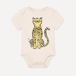 Może przedstawiać: Kremowy śpioszek dla niemowląt z kreskówkową ilustracją uśmiechniętego lamparta w kolorze żółtym i czarnym.