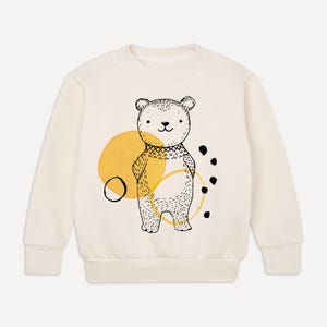 Könnte beinhalten: Ein cremefarbener Pullover mit einer schwarz-weißen Illustration eines Bären, der einen gelben Pullover trägt. Der Bär steht vor einem gelben Kreis mit schwarzen Punkten drum herum.