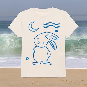 Może przedstawiać: Biała koszulka z niebieskim królikiem, księżycem, falami i małym wzorem kwiatu.