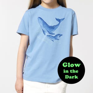 Può includere: Maglietta blu chiaro con un design di due balene che brillano al buio. La balena più grande sta saltando fuori dall'acqua e quella più piccola sta nuotando sotto. Il testo "Glow in the Dark" è stampato su un cerchio nero.