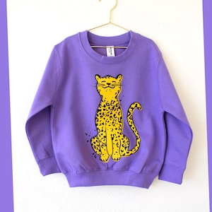Può includere: Una felpa viola con un disegno di leopardo giallo. Il leopardo è seduto con la coda arricciata. Il capo è appeso a una gruccia dorata su sfondo bianco. Il design è in stile giocoso e stravagante.