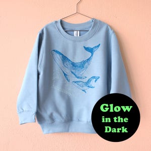 Może przedstawiać: Jasnoniebieska bluza z nadrukiem przedstawiającym niebieskiego wieloryba i jego cielę. Grafika zawiera tekst "Glow in the Dark" w czarnym okręgu. Bluza wisi na metalowym wieszaku.