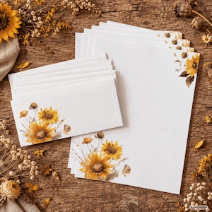 Könnte beinhalten: Ein Stapel weißes Briefpapier und passende Umschläge, jeweils verziert mit einer Aquarellillustration von gelben Sonnenblumen und getrockneten Wildblumen. Das florale Design befindet sich in der unteren linken Ecke der Umschläge und des Papiers.