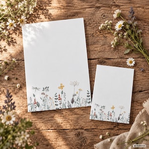 Könnte beinhalten: Zwei rechteckige weiße Briefpapierbögen mit einer zarten Aquarellillustration von Wildblumen, Gräsern und einem kleinen gelben Schmetterling am unteren Rand. Das Papier hat eine glatte Textur und liegt auf einer rustikalen Holzoberfläche.