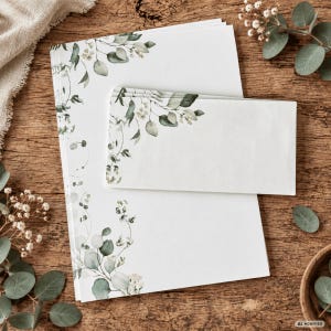 Könnte beinhalten: Ein Stapel weißes Briefpapier und ein passender Umschlag, beide verziert mit einem zarten botanischen Druck aus grünen Eukalyptusblättern und kleinen weißen Blüten an den Rändern, angeordnet auf einer strukturierten Holzoberfläche.