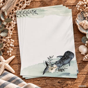 Könnte beinhalten: Ein Stapel rechteckiger Briefpapierbögen mit einer Aquarellillustration eines Walhais, der unten mit weißen Blüten und grünen Blättern verziert ist, sowie einem sanften grünen Farbauftrag oben und einem zarten Blattrand an der Oberkante.