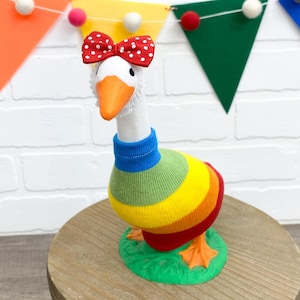 May include: A whimsical goose figurine with a white head, orange beak, and a red bow with white polka dots. The goose wears a rainbow-striped body and a blue collar. It stands on a green base, with a colorful banner in the background.
