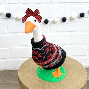 May include: A white goose figurine wearing a black, red, and gray patterned sweater and a red plaid bow. The goose stands on a green base, with orange feet and beak. The figurine is displayed on a wooden surface.