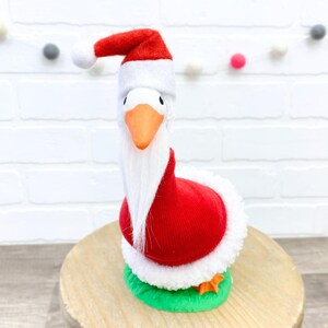 May include: A whimsical goose figurine dressed in a Santa Claus outfit. The goose wears a red and white Santa hat and a red coat trimmed with white fur. It has an orange beak and feet, and a long white beard. The figurine stands on a green base.