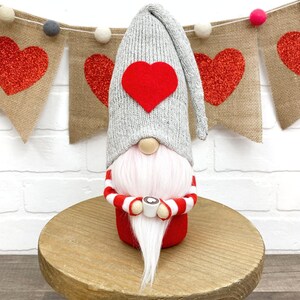 May include: A gray and red gnome holding a pink mug with a white marshmallow. The gnome is wearing a gray knit hat with a red heart on the front. The gnome is sitting on a wooden pedestal in front of a burlap banner with red glitter hearts.