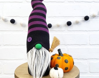 May include: A Halloween gnome with a black and purple striped hat, a white beard, and a green nose. It has a small broom and is next to an orange pumpkin with black polka dots and a small white pumpkin. The items are on a wooden stand.