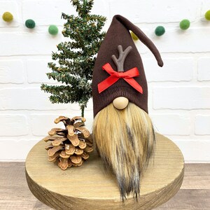 May include: A brown gnome with a brown knit hat and a red bow. The hat has a brown antler on top. The gnome has a long, brown beard and is sitting on a wooden surface.