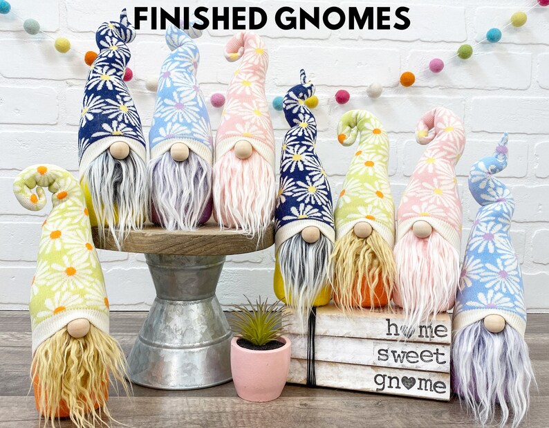 Może przedstawiać: Kolekcja ręcznie robionych gnom&oacute;w z długimi, kręconymi brodami i wysokimi kapeluszami we wzory. Kapelusze mają wz&oacute;r stokrotki w r&oacute;żnych kolorach, w tym niebieskim, r&oacute;żowym i ż&oacute;łtym. Gnomy są wystawione na drewnianym stojaku i książkach, z napisem "FINISHED GNOMES".