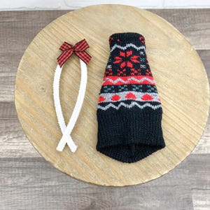 May include: A black knitted dog sweater with a red and white snowflake pattern. A white rope-like leash with a red and black plaid bow is next to the sweater. The items are on a light brown wooden surface.