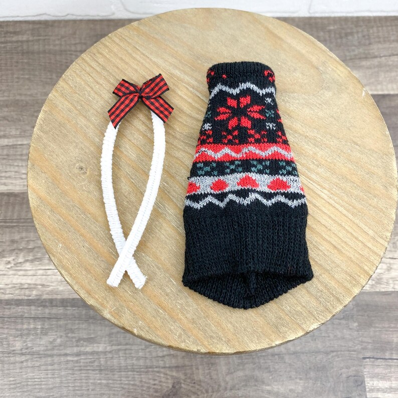 May include: A black knitted dog sweater with a red and white snowflake pattern. A white rope-like leash with a red and black plaid bow is next to the sweater. The items are on a light brown wooden surface.