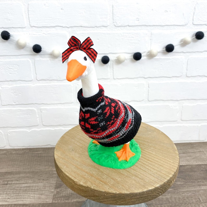 May include: A white goose figurine wearing a black, red, and gray patterned sweater and a red plaid bow. The goose stands on a green base, with orange feet and beak. The figurine is displayed on a wooden surface.