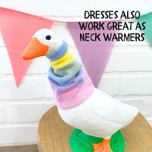 May include: A white goose figurine wearing a colorful striped neck warmer. The goose has an orange beak and feet, and is perched on a wooden surface. The text "Dresses also work great as neck warmers" is displayed above the goose.