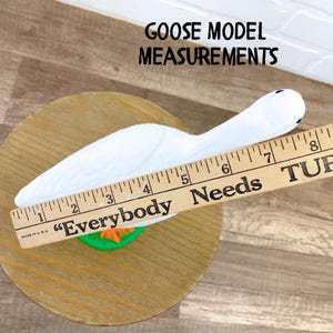 May include: A white goose model with a wooden ruler showing measurements. The goose is on a wooden base with a green and orange accent. The ruler reads "Everybody Needs". The text "GOOSE MODEL MEASUREMENTS" is at the top.