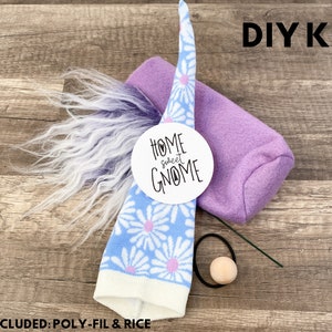Może przedstawiać: Zestaw DIY do wykonania gnoma z niebiesko-białym kapeluszem w stokrotki, fioletowym korpusem, biało-fioletową brodą z imitacji futra i drewnianym nosem. Zestaw zawiera okrągłą etykietę z napisem "Home Sweet Gnome". Widoczny jest tekst "DIY KIT".