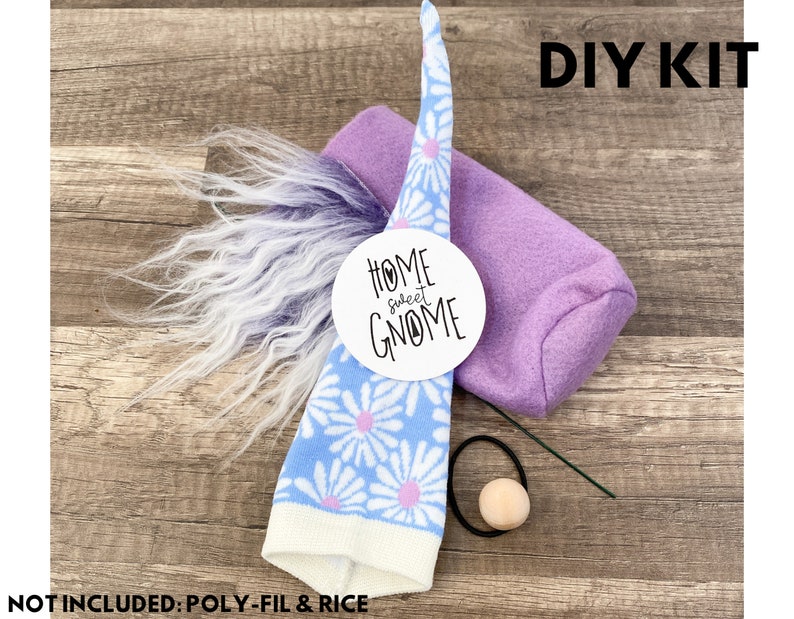 Może przedstawiać: Zestaw DIY do wykonania gnoma z niebiesko-białym kapeluszem w stokrotki, fioletowym korpusem, biało-fioletową brodą z imitacji futra i drewnianym nosem. Zestaw zawiera okrągłą etykietę z napisem "Home Sweet Gnome". Widoczny jest tekst "DIY KIT".