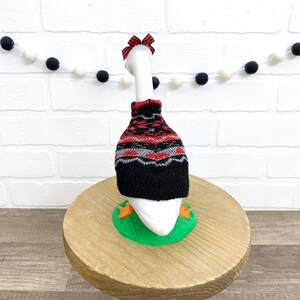 May include: A white goose figurine wearing a black, red, and gray patterned sweater and a red bow. The goose stands on a green base with orange feet. The background includes a white brick wall and a garland of black and white felt balls.