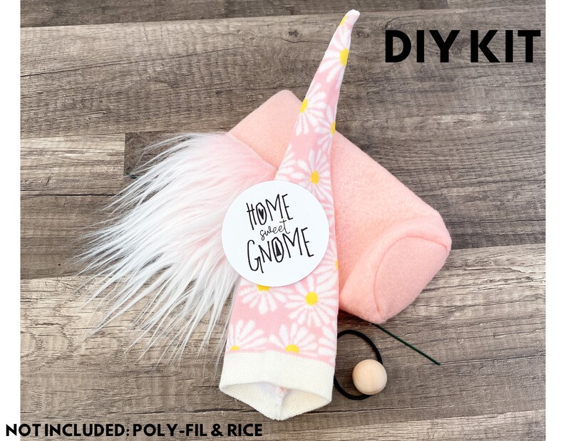 Może przedstawiać: Zestaw DIY do wykonania gnoma. Zestaw zawiera r&oacute;żowy filcowy korpus, czapkę we wz&oacute;r białych stokrotek na r&oacute;żowym tle, białą brodę z imitacji futra, drewnianą koralik i czarną gumkę. W zestawie znajduje się r&oacute;wnież okrągła etykieta z napisem "HOME sweet GNOME".