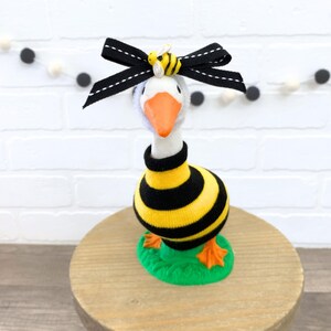 May include: A white goose figurine wearing a black and yellow striped sweater and a black bow with a yellow bee on top. The goose is standing on a green base.