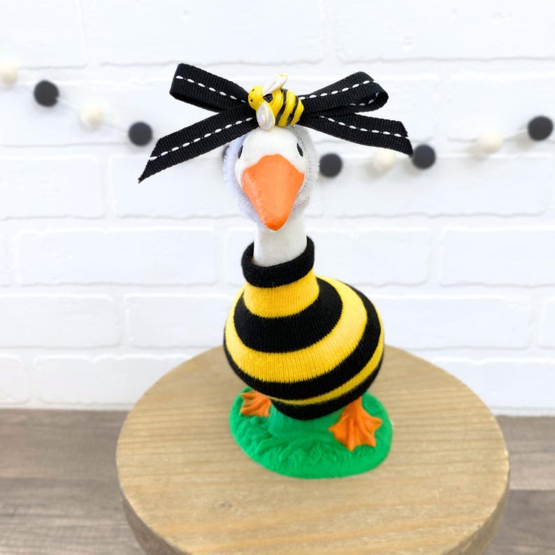 May include: A white goose figurine wearing a black and yellow striped sweater and a black bow with a yellow bee on top. The goose is standing on a green base.