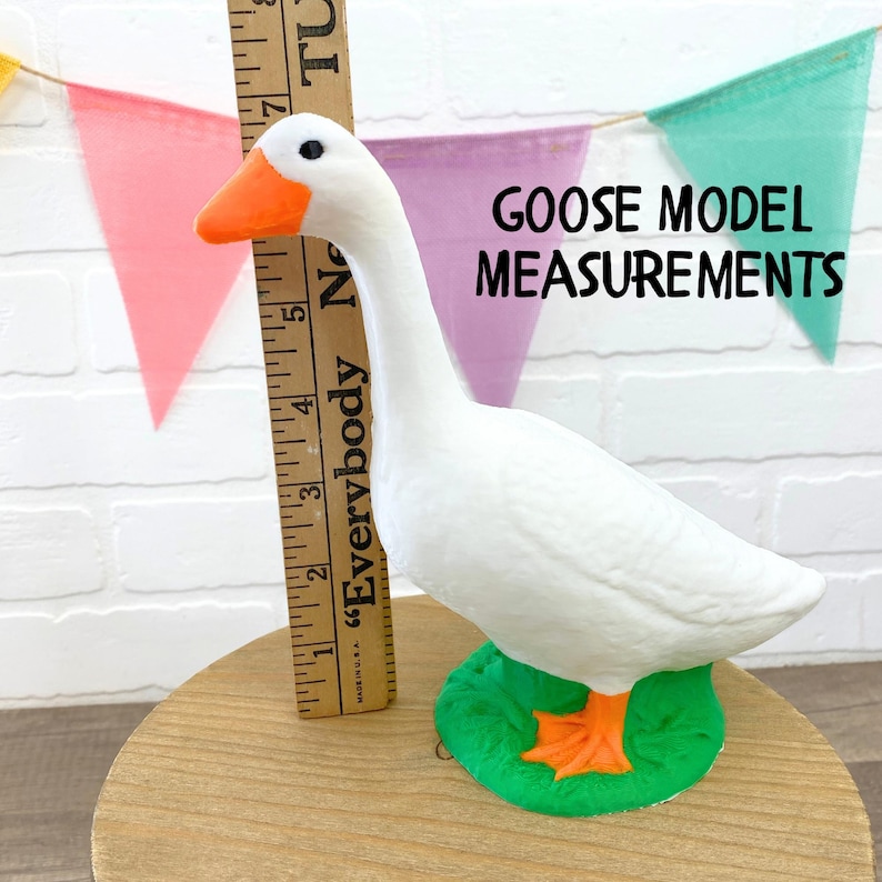 May include: A white goose model with an orange beak and feet, standing on a green base. A wooden ruler is placed next to the goose. The text "GOOSE MODEL MEASUREMENTS" is displayed. Colorful pennants are in the background.