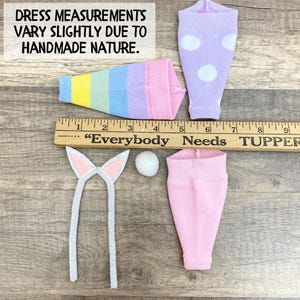 May include: A collection of handmade doll accessories, including a rainbow-striped cone, two pairs of socks, one lavender with white polka dots, and one pink. Also included are a pair of bunny ears and a small white pom-pom. The text in the image reads: "DRESS MEASUREMENTS VARY SLIGHTLY DUE TO HANDMADE NATURE."