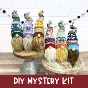 Puede incluir: Seis gnomos decorativos con sombreros estampados y largas barbas se exhiben en un soporte de madera. Los sombreros presentan varios colores y diseños, incluyendo copos de nieve y patrones geométricos. El texto "DIY MYSTERY KIT" está en la parte inferior.