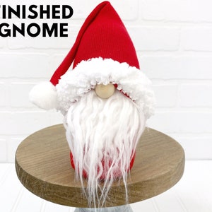 Puede incluir: Un gnomo rojo y blanco terminado con una barba blanca y un pomp&oacute;n blanco en la parte superior del gorro. El gnomo est&aacute; sentado en un pedestal de madera.