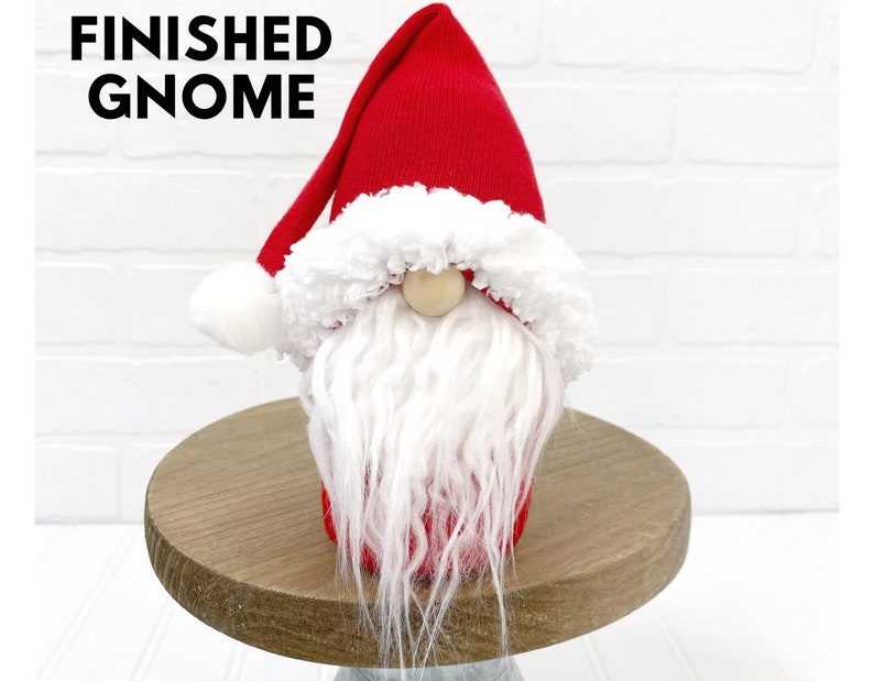 Puede incluir: Un gnomo rojo y blanco terminado con una barba blanca y un pomp&oacute;n blanco en la parte superior del gorro. El gnomo est&aacute; sentado en un pedestal de madera.