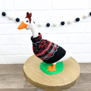 May include: A white goose figurine wearing a black, red, and gray patterned sweater and a red and black striped headband. The goose has an orange beak and feet and is standing on a green base atop a wooden stand.