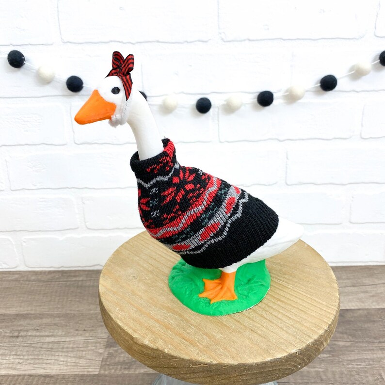 May include: A white goose figurine wearing a black, red, and gray patterned sweater and a red and black striped headband. The goose has an orange beak and feet and is standing on a green base atop a wooden stand.