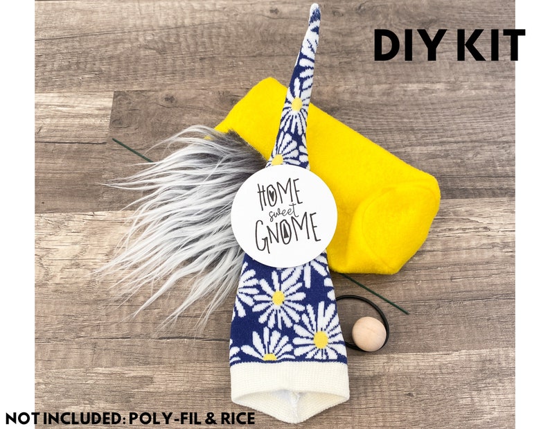 Może przedstawiać: Zestaw DIY do wykonania gnoma. Zestaw zawiera niebiesko-białą czapkę w stokrotki, ż&oacute;łty filcowy korpus, szare sztuczne futro, drewnianą kulkę i czarną gumkę. Biała etykieta głosi "Home Sweet Gnome". Widoczny jest r&oacute;wnież tekst "DIY KIT".