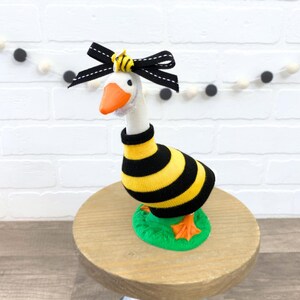 May include: A white goose figurine wearing a black and yellow striped sock, a black bow with a bee charm, and standing on a green base.