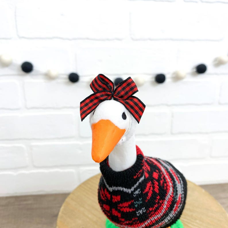 May include: A white goose figurine with an orange beak and a red and black bow on its head. The goose wears a black, red, and gray patterned sweater. The figurine is on a wooden surface.