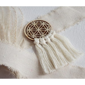 Könnte beinhalten: Ein Holz-Kreis mit einem geometrischen Muster und einem weißen Quasten, der daran hängt. Der Kreis liegt auf einem weißen Stoffhintergrund.