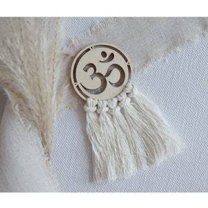 Könnte beinhalten: Ein Holzkreis mit einem geschnitzten Om-Symbol und einem weißen Quastenfransen. Der Kreis befindet sich auf einem weißen Stoffhintergrund.