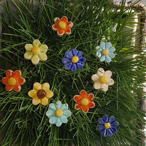 Könnte beinhalten: Sortiment an bunten Keramik-Blumennadeln in verschiedenen Blautönen, Gelb, Rot und Weiß. Die Nadeln sind auf einem Bett aus grünem Gras angeordnet.