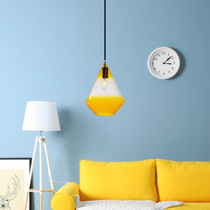 May include: A yellow sofa with a white throw blanket in a living room with a yellow and white glass pendant light, a white clock, and framed artwork on the wall.