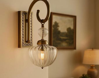 May include: A wall-mounted light fixture featuring a rectangular brass backplate with a lattice pattern, a curved brass arm, and a clear, ribbed glass shade. The fixture holds a single glowing bulb, creating a warm, focused light effect.