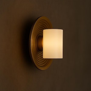 May include: A wall sconce with a circular, textured brass backplate and a cylindrical, frosted glass shade. The light fixture emits a warm, inviting glow, perfect for ambient lighting in a home or commercial space.
