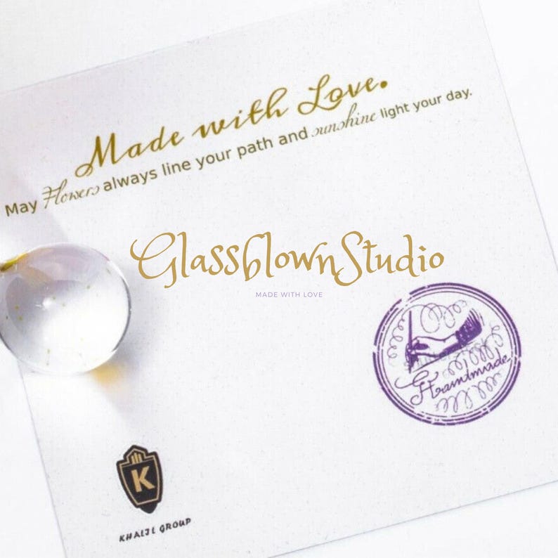 May include: A white card with gold text that reads "Made with Love" and "Glassblown Studio." A clear glass sphere is in the upper left corner. A purple stamp with the words "Handmade" and a logo is on the right.